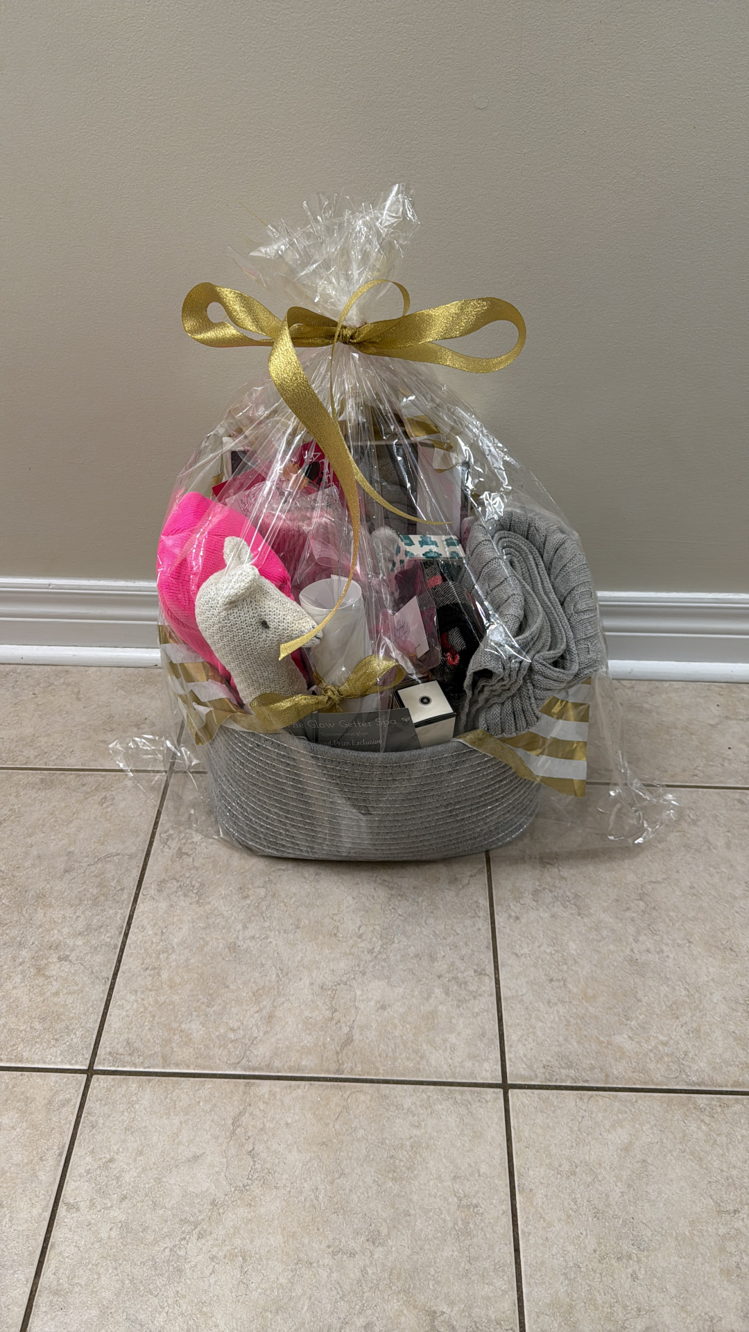 Gift basket with a pink hat, knitted toy, and spa items tied with gold ribbon