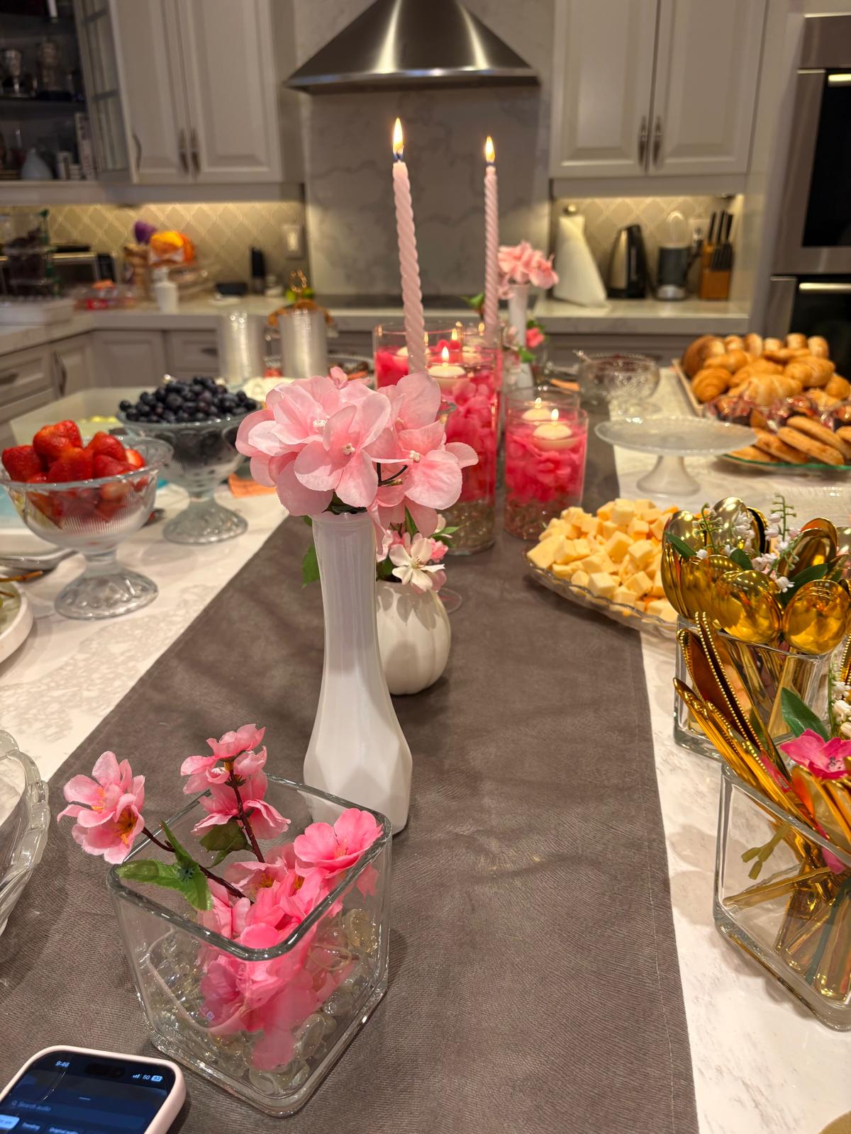 Cheese, fruit, and gold cutlery arranged on a kitchen island with pink flowers and lit candles
