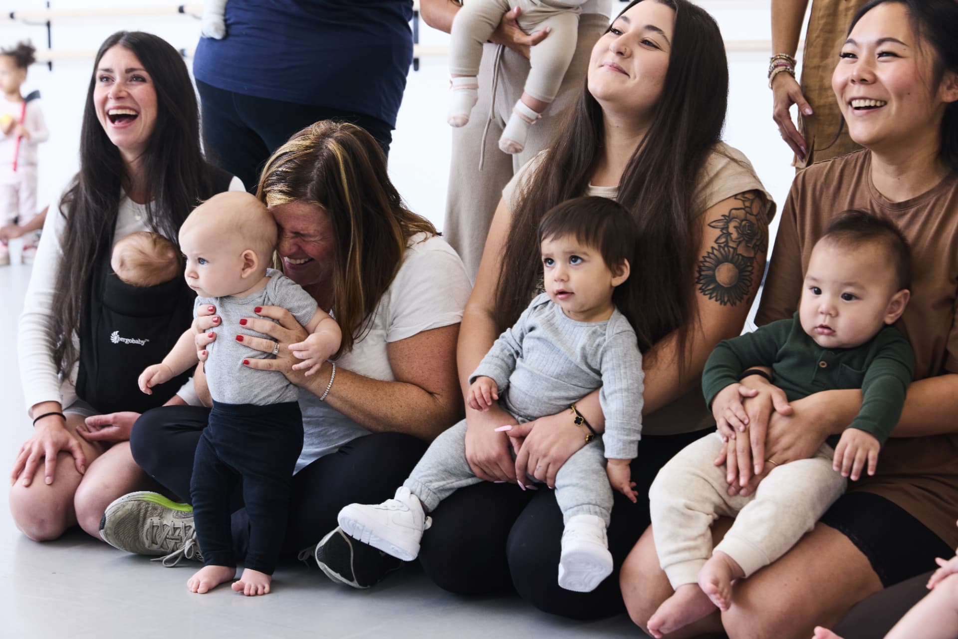 Moms sitting together on the studio floor laughing as their babies play in front of them