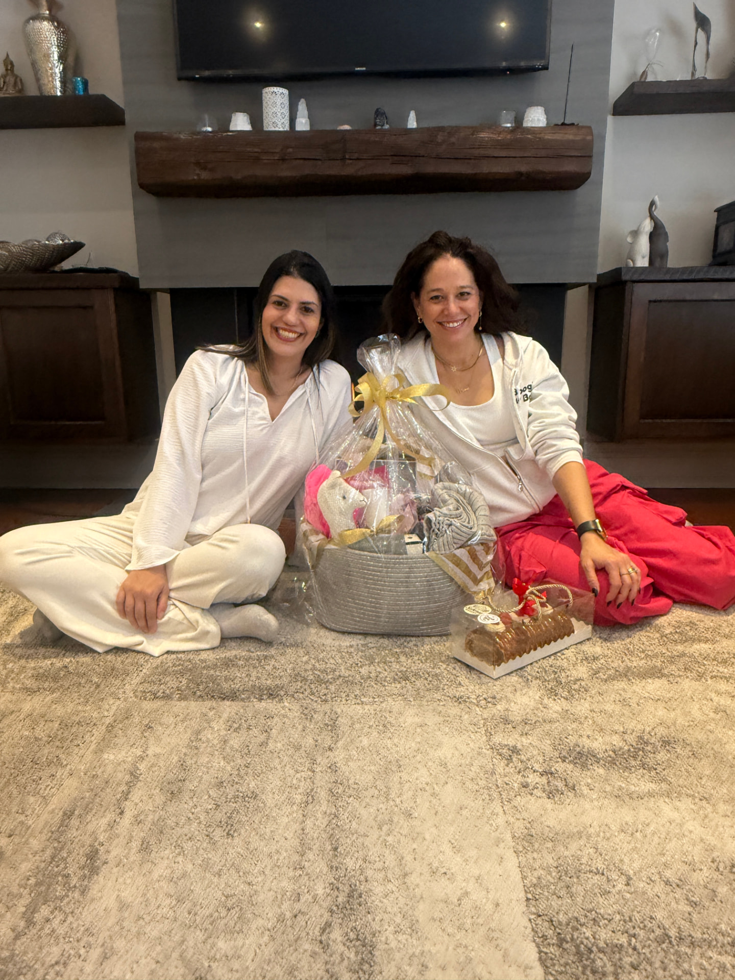 Two smiling women seated on the floor beside a wrapped gift basket and a boxed dessert in front of a fireplace