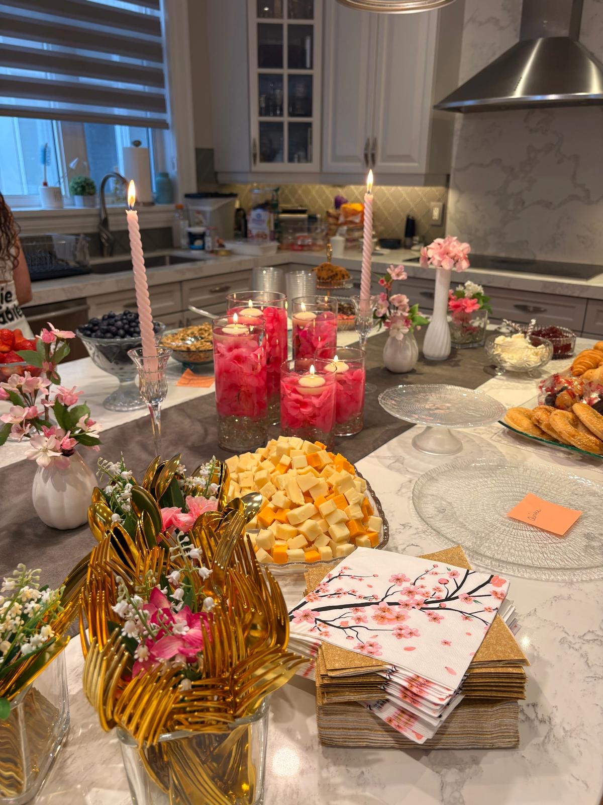 Kitchen island laid out with cubed cheese, pastries, fruit, pink candles, and gold cutlery