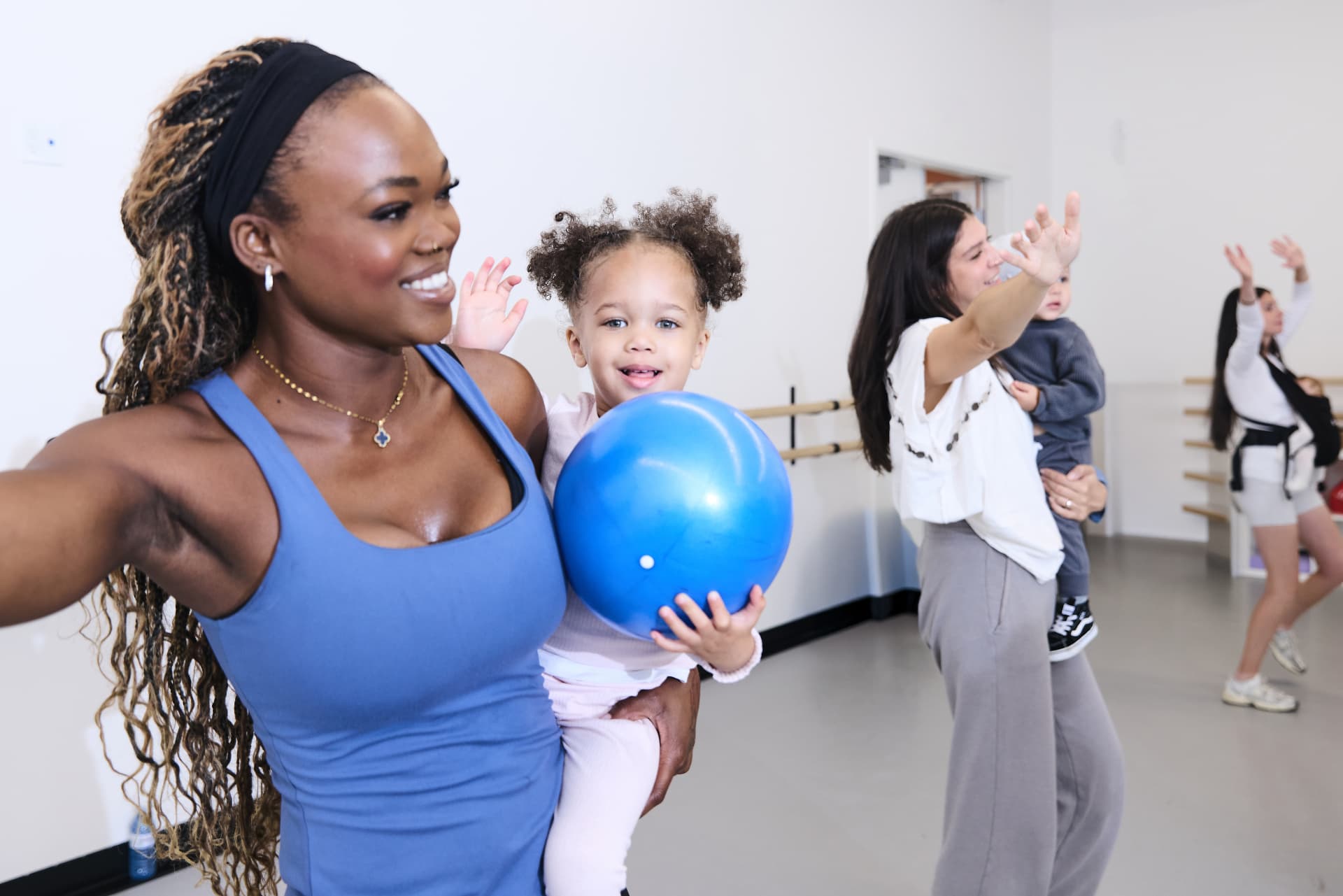 Smiling mom carrying her toddler with a blue exercise ball while other moms and babies join class behind her
